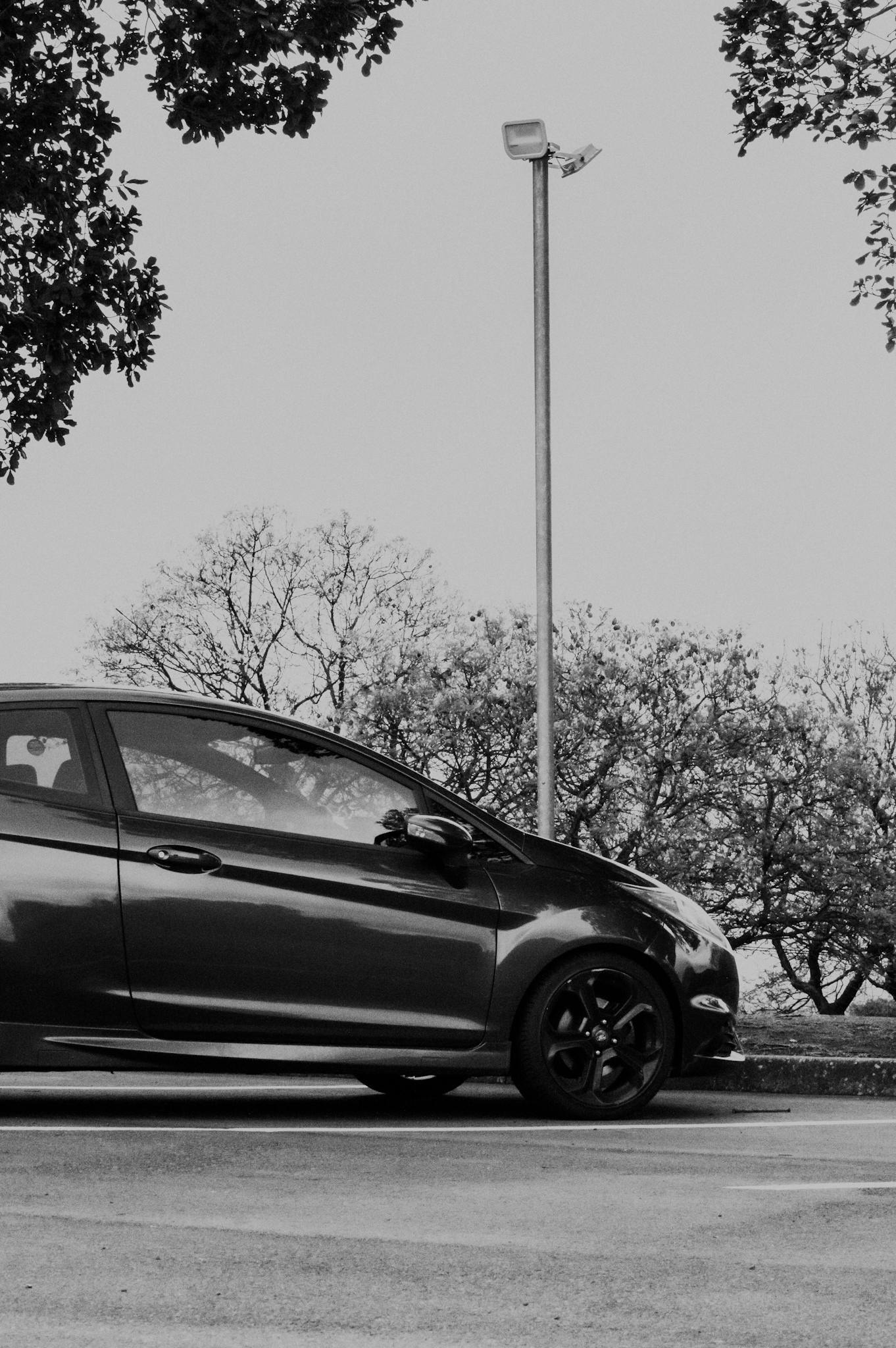 Monochrome photo of a parked car outdoors with trees in the background.